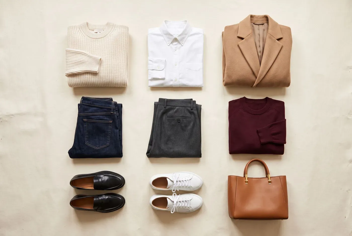 Capsule wardrobe flat lay with a cream knit, white shirt, camel blazer, jeans, trousers, burgundy knit, loafers, sneakers and a tan bag