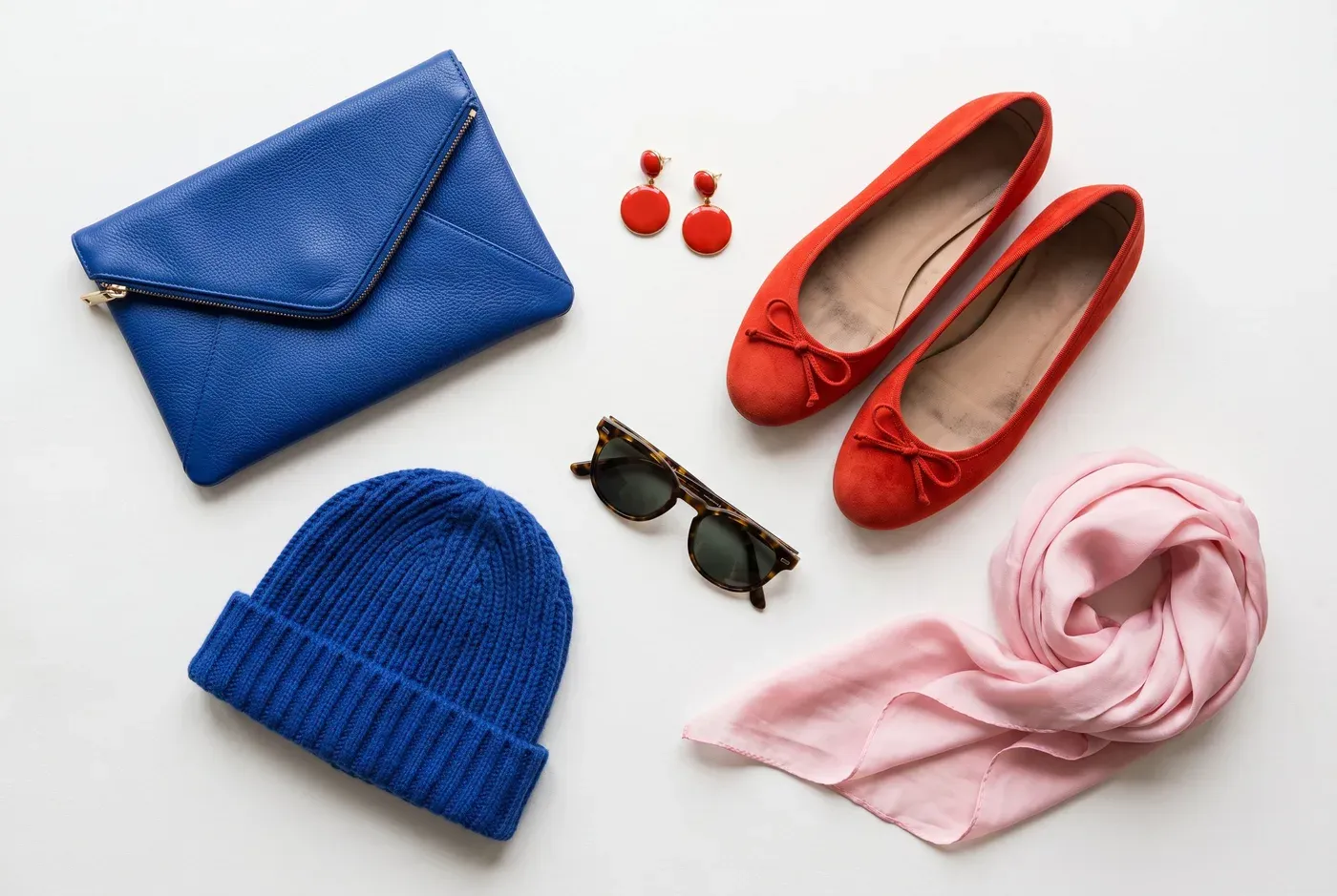 Flat lay com clutch azul cobalto, sapatilhas vermelho-tomate, gorro azul cobalto, brincos vermelhos, lenço de seda rosa e óculos de sol