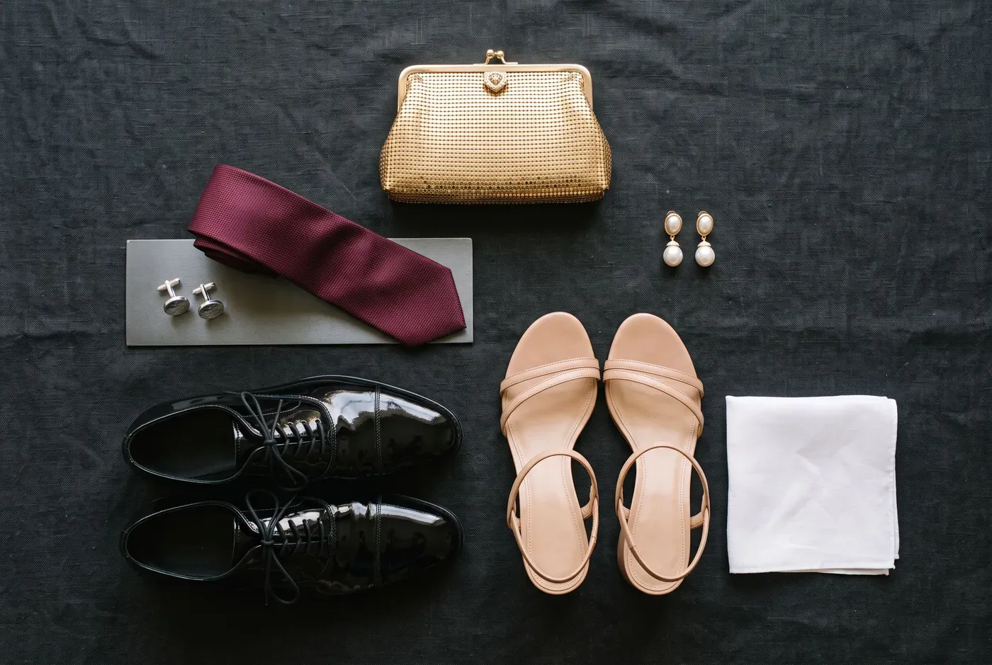 Flat lay on dark linen of a small gold clutch, burgundy silk tie, silver cufflinks, pearl drop earrings, black patent oxfords, nude block-heel sandals and a white pocket square