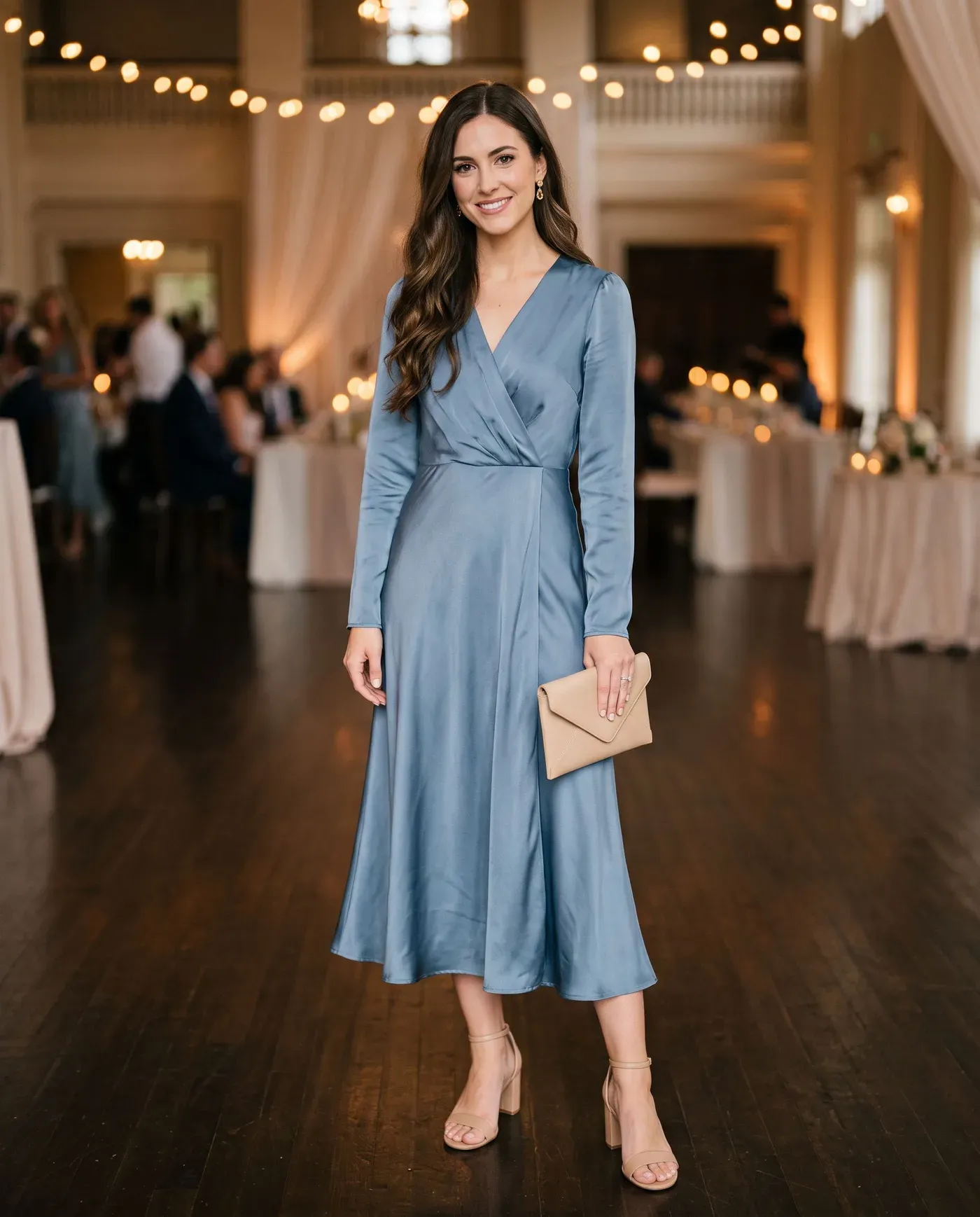 Woman in a long-sleeved dusty blue satin midi dress with nude block-heel sandals and drop earrings, holding a small clutch in a softly lit event hall