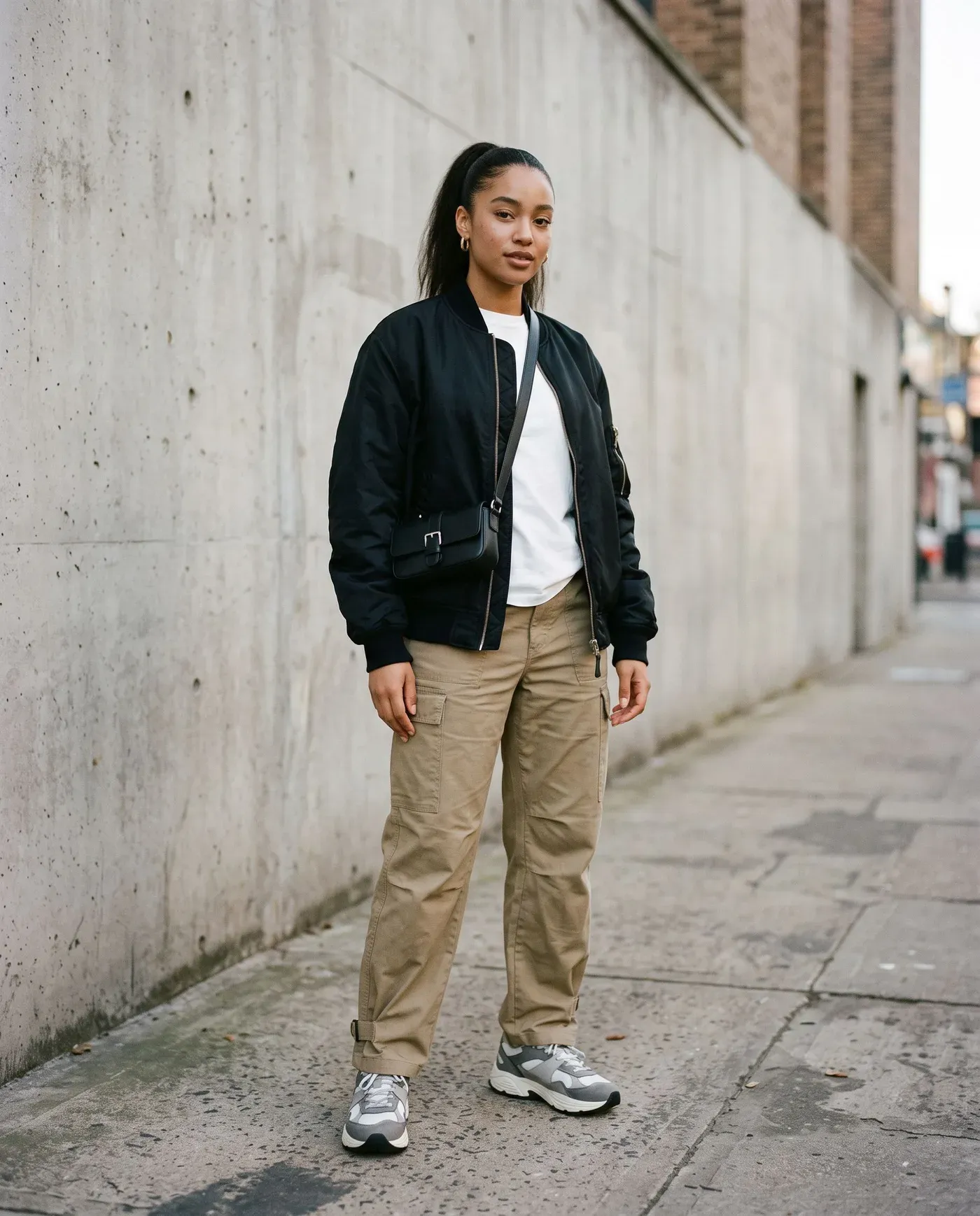 Young woman in an oversized black bomber jacket over a white T-shirt with loose khaki cargo trousers, chunky grey sneakers and a small crossbody bag