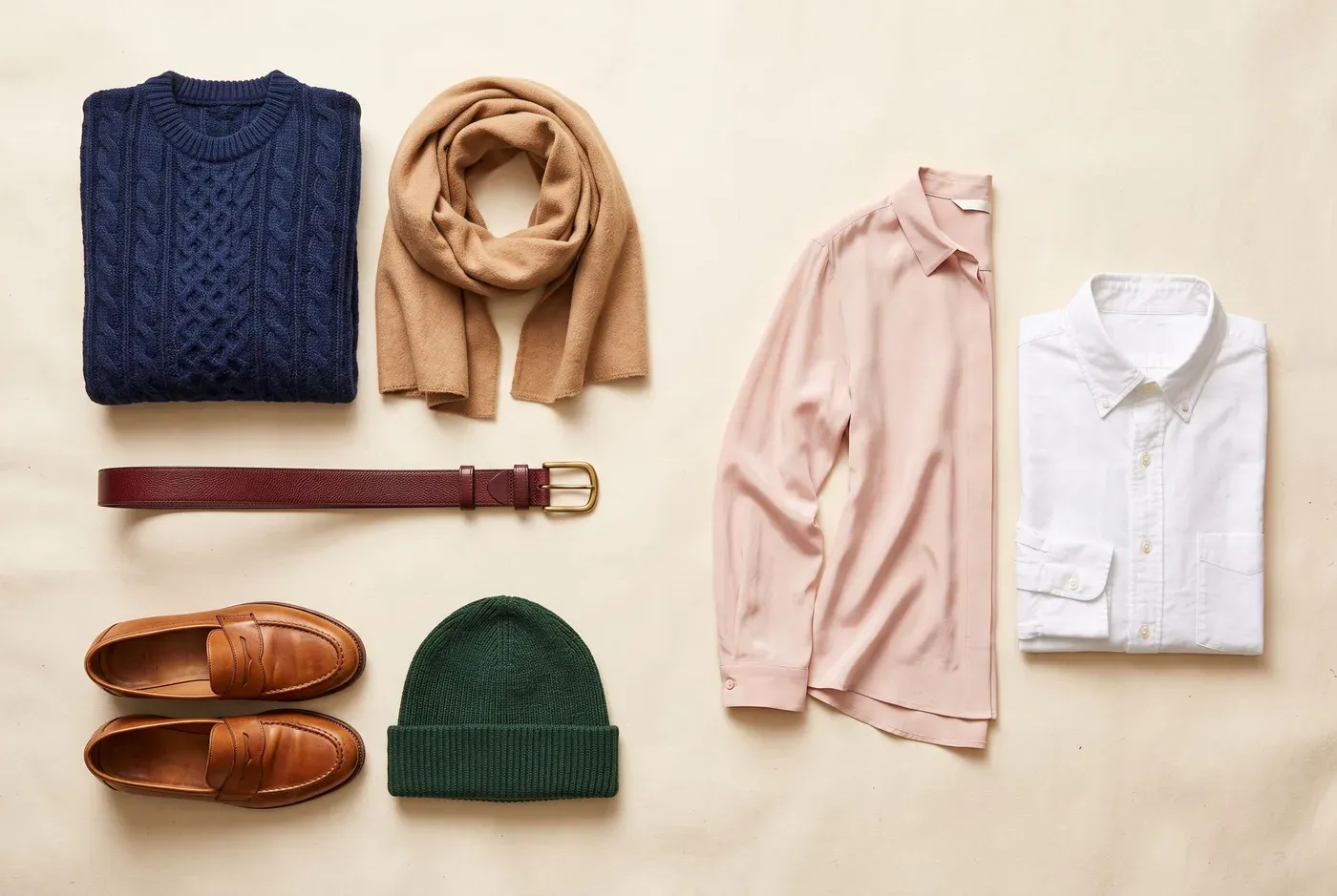 Flatlay van een donkerblauwe trui met een camel sjaal, bordeaux riem, lichtroze zijden blouse, wit oxford-overhemd, cognackleurige loafers en een bosgroene muts