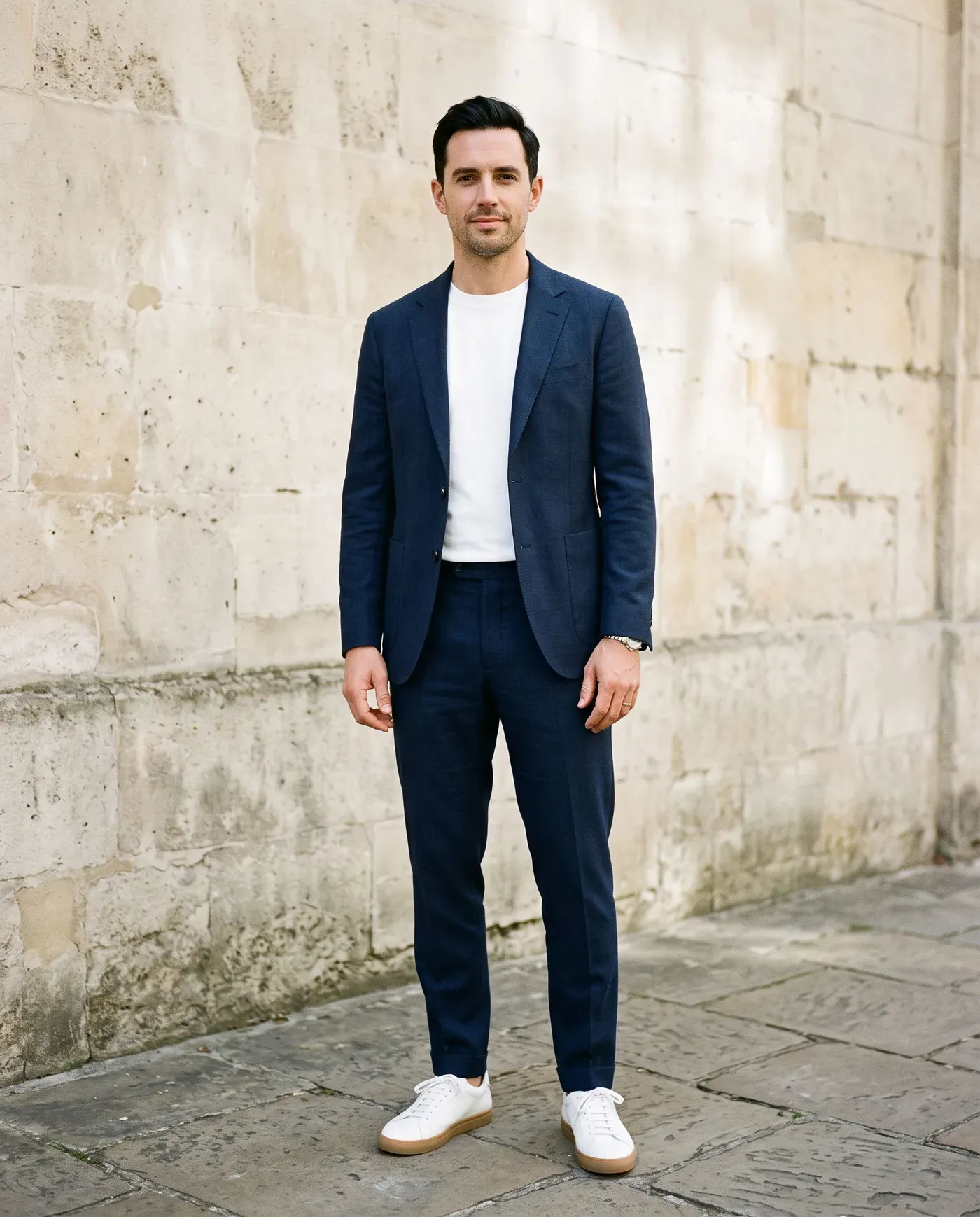 Man in een relaxed donkerblauw pak over een effen wit T-shirt met witte leren sneakers tegen een lichte stenen muur